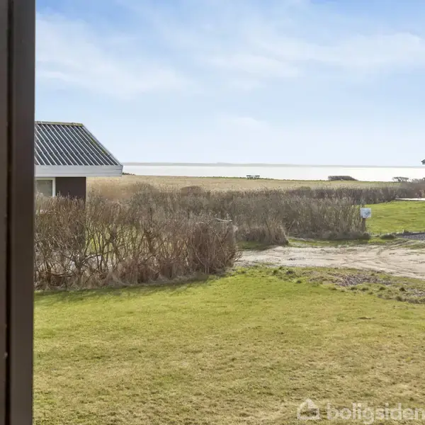 Huse står stille med græsplæne og buske i forgrunden, omgivet af sand og kystlinje, med en klar blå himmel i baggrunden.
