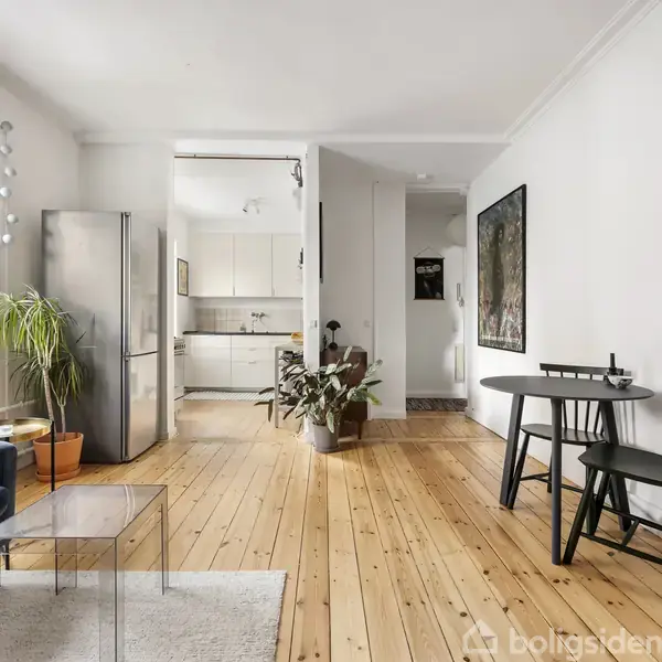 Stue med trægulv og sofa ved vindue. Plante ved siden af sofaen. Lille spisebord med to stole til højre. Køkken i baggrunden med hvidt interiør.