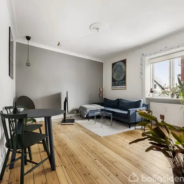 Stue med blå sofa i hjørnet, sofabord, og indendørs planter. Et rundt bord med to stole ved venstre væg. Trægulv og stort vindue giver lysindfald.