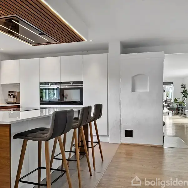 Køkkenområde med hvidt køkkenbord, tre høje barstole, og træloft. Moderne apparater indbygget i skabene. I baggrunden ses en stue med lyse trægulve og naturligt lysindfald.