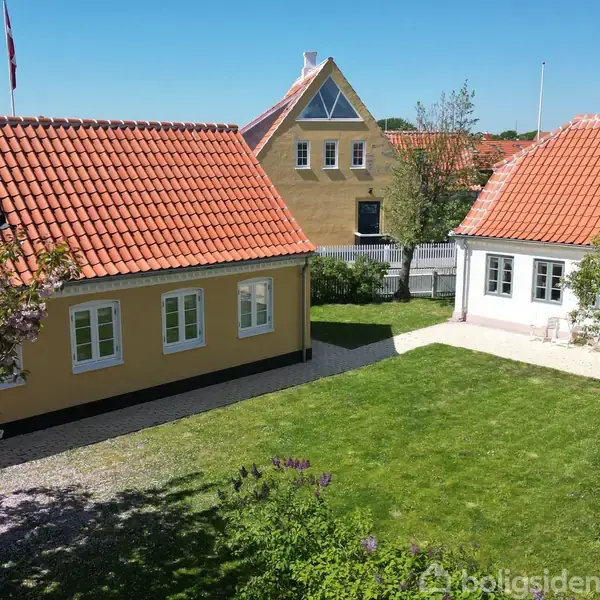 To gule huse med røde tegltage ligger i en have med grøn græsplæne og blomstrende træer. Et flag vajer ved siden af husene under en klar blå himmel.