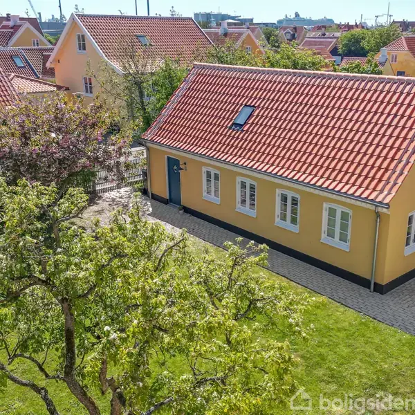 Et gult hus med rødt tegltag står i en have med frodige træer. Andre lignende huse omringer stedet i et boligområde.
