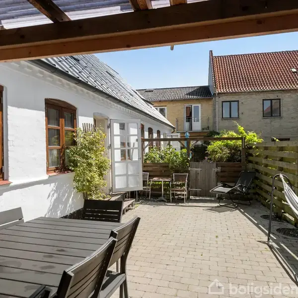 Træbord med stole står på en brolagt terrasse ved siden af et hvidt hus. En åben dør fører ind, og plantekasser pynter terrassen under klar blå himmel.