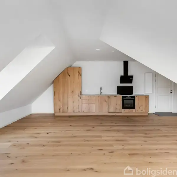 Loftlejlighed med skråvægge og trægulv. Et åbent, lyst rum med et moderne køkken i træ, sort komfur og ventilator. Intet tekst på væggene.
