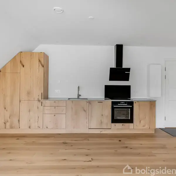 Et moderne køkken med trælåger og indbygget ovn står langs en hvid væg. Trægulv dominerer rummet, ved siden af en hvid dør og en mørk måtte.