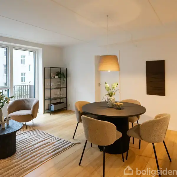 Stue med rundt spisebord omgivet af stole under en hængelampe, tæt på en stor, lys vindueparti. Elegant reol og sofabord med blomster placeret på træplankegulv.
