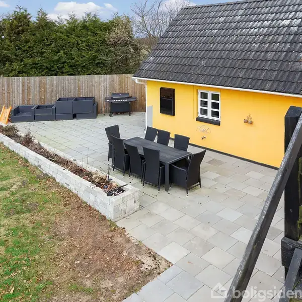 Et gult hus med sort tag ligger ved siden af en flisebelagt terrasse. På terrassen står et bord med seks sorte stole og en loungesofa. En græsplæne grænser op til terrassen.