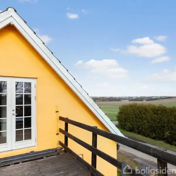 Et gult hus med en hvid terrassedør åbner mod en træveranda. I baggrunden ses en grøn mark og en hæk under en blå himmel med skyer.