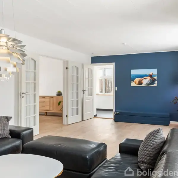 En moderne stue med sorte lædersofaer og et rundt, hvidt sofabord. En unik loftslampe hænger over bordet, og i baggrunden ses en åben dør og en blå væg.