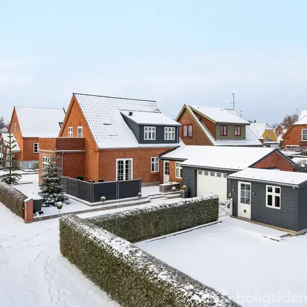 Et rødt murstenshus med snedækket tag og snedækket have er omgivet af en grøn hæk. Flere lignende huse og et skur ses i det snedækkede nabolag.