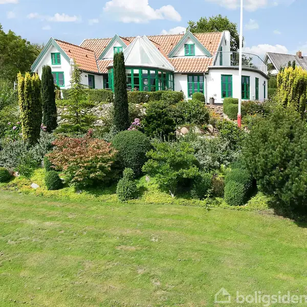 Et stort hus med rødt tegltag og grønne skodder står omgivet af en velplejet have med forskellige buske og træer. Skyfri blå himmel danner baggrund.