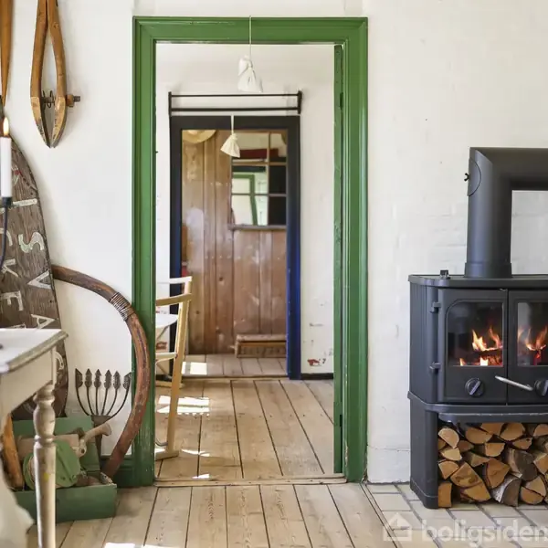 Brændeovn står i et lyst rum, fyret op med flammer indeni. Brændestakke ved siden af, og dør med grøn ramme fører til et andet værelse. Hyggelig, rustik atmosfære.
