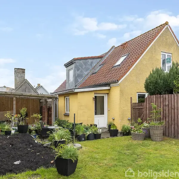 Gul murstenshus med rødt tag står i en have med grøn græsplæne og planter i krukker. En træhegn omkranser ejendommen. Skyfri blå himmel i baggrunden.