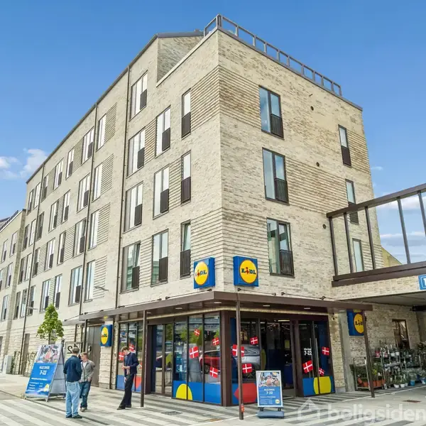 Supermarkedbygning med Lidl-logo, personer går ind gennem indgangen. Omgivet af parkeringsplads med skilt, der viser "70 Gratis P-pladser". Moderne lejlighedsbygninger i baggrunden, klar himmel.