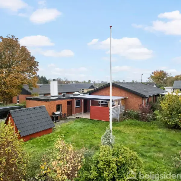 Et rødt murstenshus med fladt tag står i en have med græsplæne og buske. Et flagstang er placeret midt i plænen. Naboer og træer ses i baggrunden.