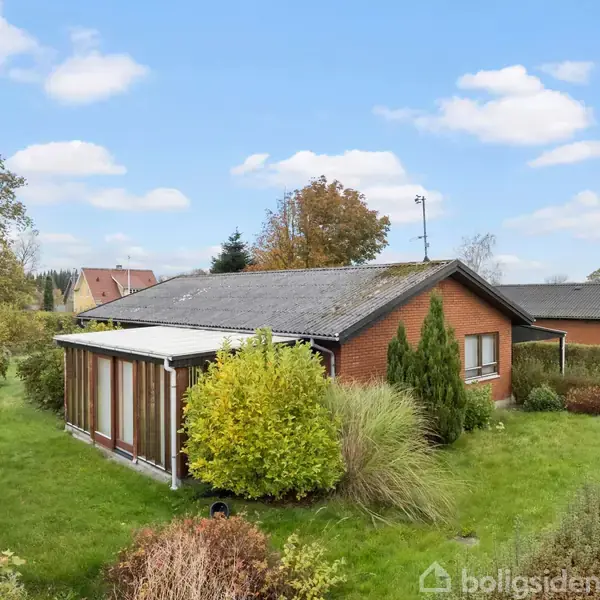 Et rødt murstenshus med en stor have omringet af buske og træer. Huset har en tilbygning med glasparti. Klart himmel med enkelte skyer i baggrunden.