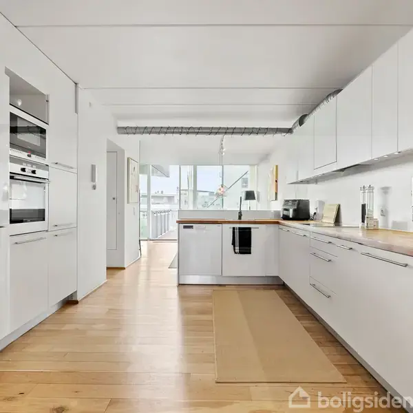 Et moderne køkken med hvide skabe og træbordplader. En ovn er integreret i et skab. Der er træparketgulve og et åbent vindue som leder til en balkon med udsigt.