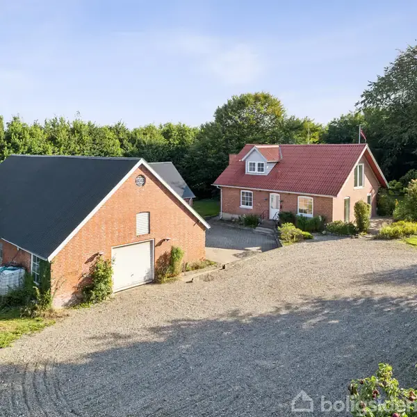 Et rødt murstenshus med hvidt træværk og en rødtag. Der står en garagebygning med sort tag til venstre. Omgivet af en grusplads og grønne træer.