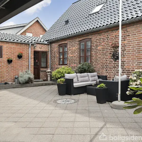 En gårdhave med moderne møbler står stille i en flisebelagt gårdsplads omkranset af røde murstenshuse med tagvinduer og plantekrukker, som skaber en hyggelig atmosfære.