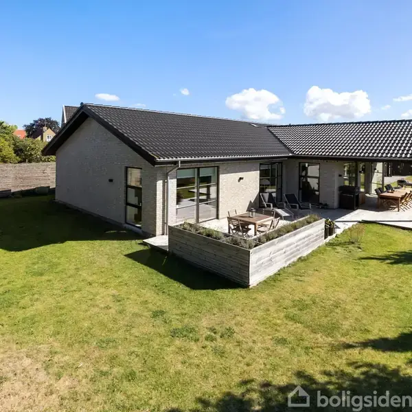 En moderne villa med fladt tag ligger i en solrig have. Terrassen er udsmykket med havemøbler. Blå himmel og enkelte skyer danner en rolig baggrund.