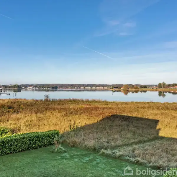 En skygge fra et hus falder på en græsmark ved siden af en stille sø. Landskabet er roligt med en klar blå himmel og nogle fjerne træer langs horisonten.