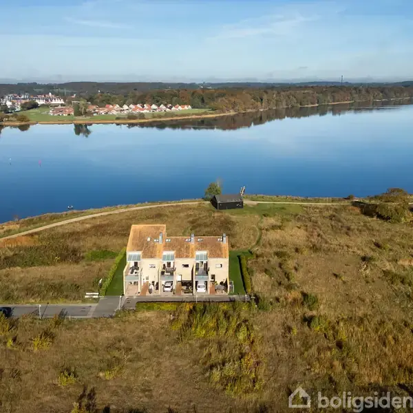 En række huse med fladt tag står i en åben natur ved en stor sø. I baggrunden ses en by på den modsatte bred.