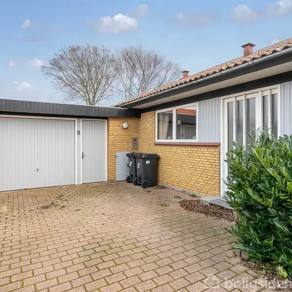 Garage med hvid dør og nummer 5 står på belagt indkørsel ved gul murstenshus. To skraldespande ved siden af garagen. Grønne buske står foran huset. Himmel med let skydække.