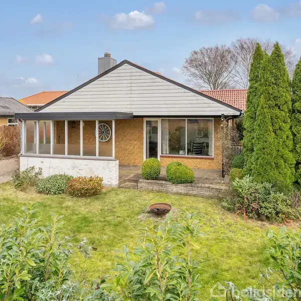 Et enkelt enfamiliehus med gul murstensvæg og skråt tag står i en frodig have med buske og træer. Stor vinduesparti i huset vender mod haven.