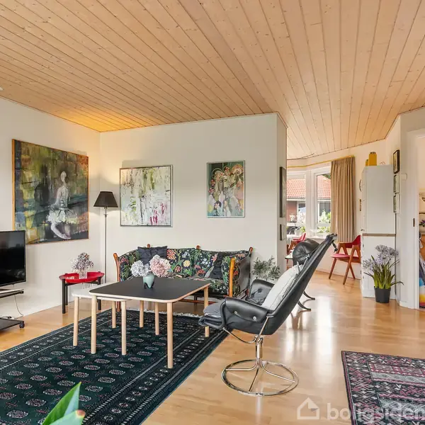 Stue med sofagruppe og tv, dekoreret med farverige malerier. Rummet har trægulv og træloft, bordlampe ved sofaen. Bagved er åben adgang til spisestue med bord og stole.