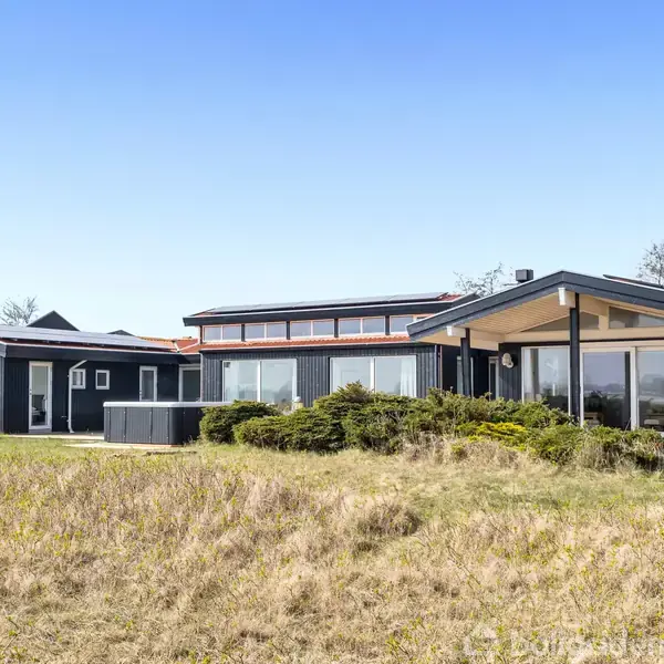 Et moderne sommerhus med mørke træbeklædninger står roligt i et åbent område med græs og buske under en klar blå himmel. Store vinduer reflekterer omgivelserne.