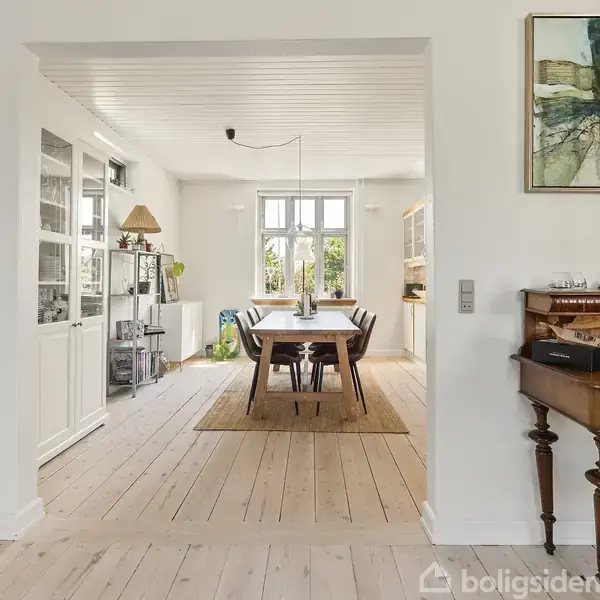 Spisestue med træbord og stole står på et tæppe i et lyst rum med trægulv. Der er store vinduer, en reol til venstre, og et klaver til højre.