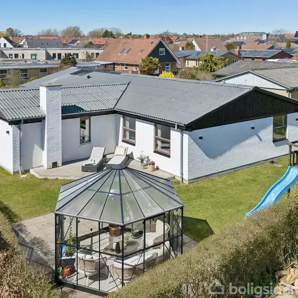 Et hvidt murstenshus med sort tag står omgivet af en velplejet have. Foran er en glaspavillon og en blå rutsjebane. Huse og veje ses i baggrunden.