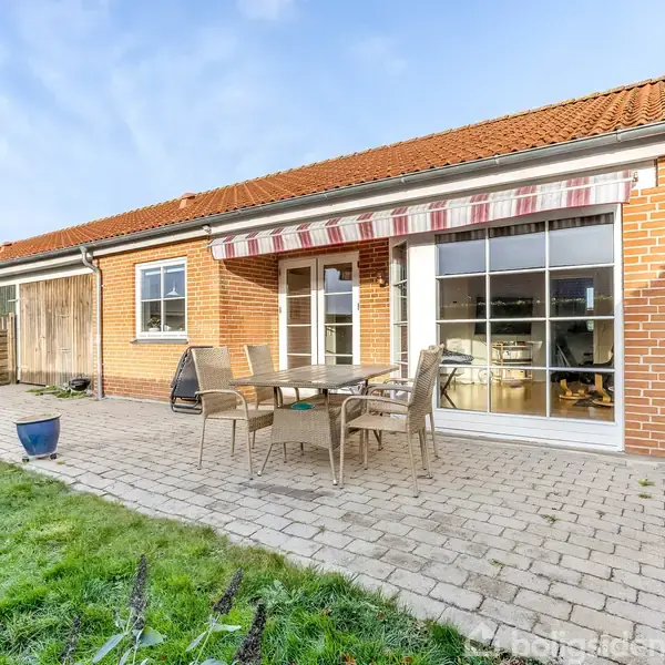 Et rødt murstenshus med en terrasse, hvor der står et bord og stole. Terrassen har en stribet markise og er omgivet af en grøn græsplæne.