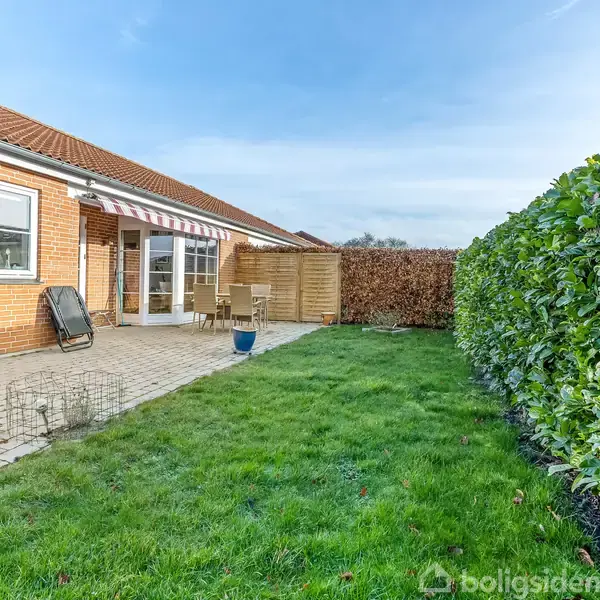 Hus med rødt tegltag og store vinduer står ved en terrasse med fliser. Grøn græsplæne omgivet af en tæt hæk. Klapstol og bord på terrassen.