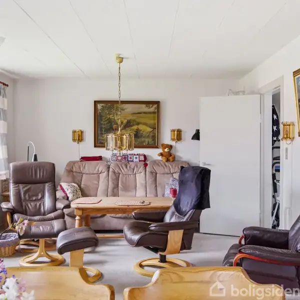 Brune lænestole står foran en beige sofa i en stue. Et sofabord er i midten med malerier på væggene. En dør til højre fører til et andet rum.