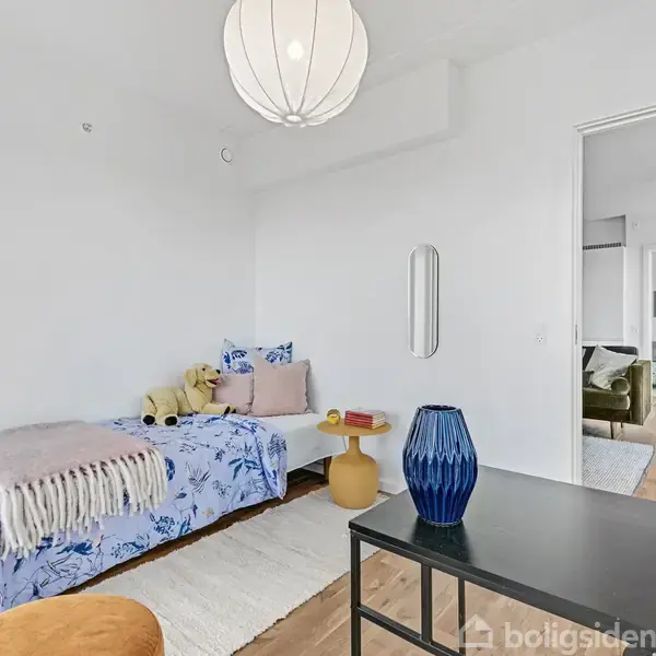 Et lille værelse med en enkeltseng dækket af et blomstret sengetæppe, placeret ved siden af en guld vase på bordet. Et åbent dørhul leder til stue med sofa.