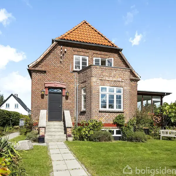 Rødt murstenshus står stille med rødt tegltag og hvide vinduer, omgivet af velholdt have med sti og grønne planter, under en klar blå himmel.