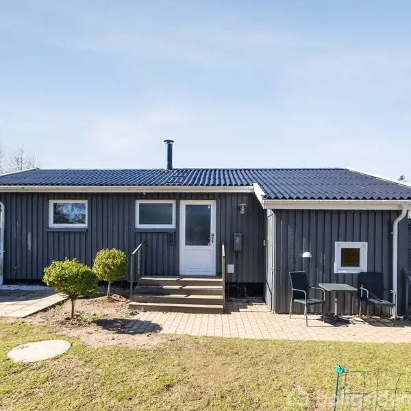 Et enkelt gråt sommerhus står stille i en have med en brolagt sti foran. En campingvogn er parkeret til venstre, og der er havemøbler på en terrasse til højre.