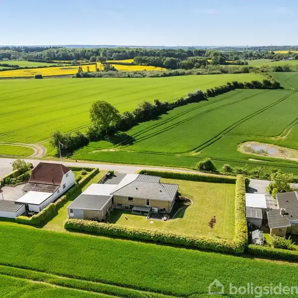 Huse står stille blandt velplejede græsplæner og hække i en landlig dal med åbne marker og skove i baggrunden. Sive mindre bygninger ses ved en vej.