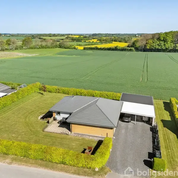 Et hus med gråt tag og carport ses fra oven. Det ligger i en velplejet have med hække, omgivet af store grønne marker og enkelte træer i baggrunden.