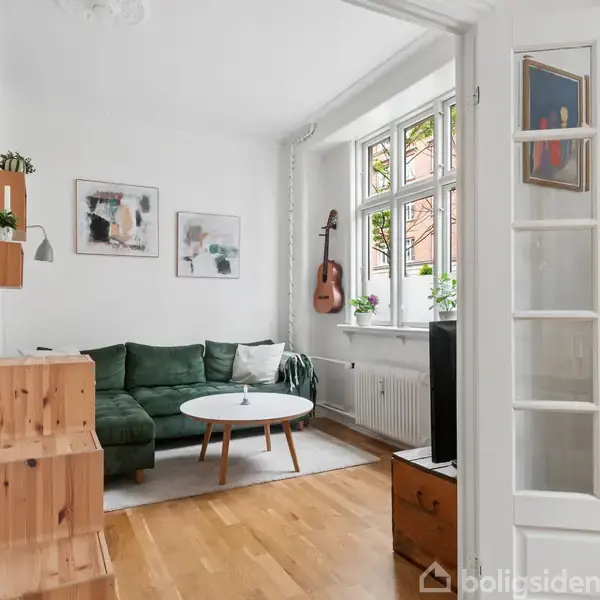 Grøn sofa står i lys stue med hvid væg, træmøbler og runde borde, mens en guitar hænger ved vinduet. Malerier og planter dekorerer rummet. Ingen tekst til stede.