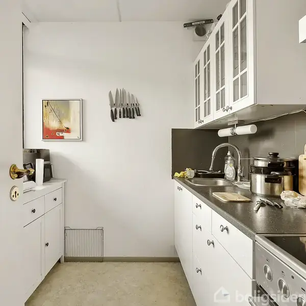 Et lille køkken har hvide skabe og en sort bordplade. Der hænger knive på væggen ved siden af et billede. Køkkenet er velorganiseret med forskellige køkkenredskaber.