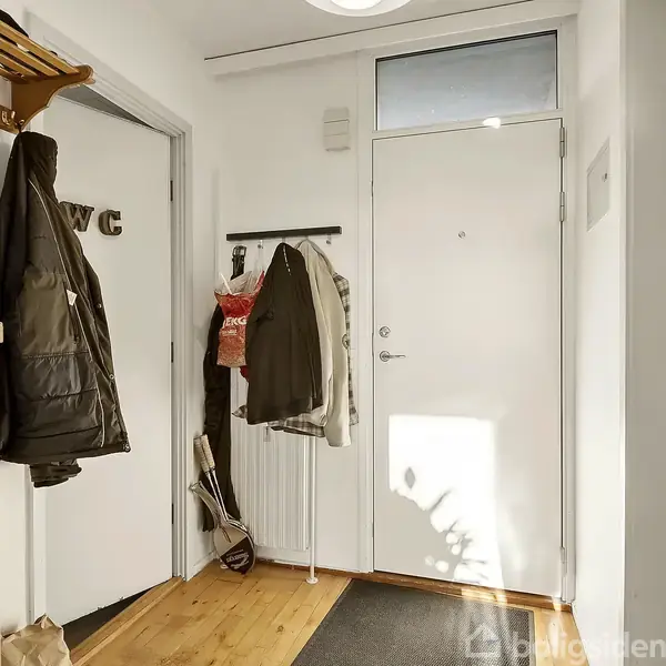 Dør med påhængt tøj, åbner mod en lys gang. Venstre side har kroge med jakker. En dør til venstre viser teksten "WC". Trægulv og hvidmalede vægge.