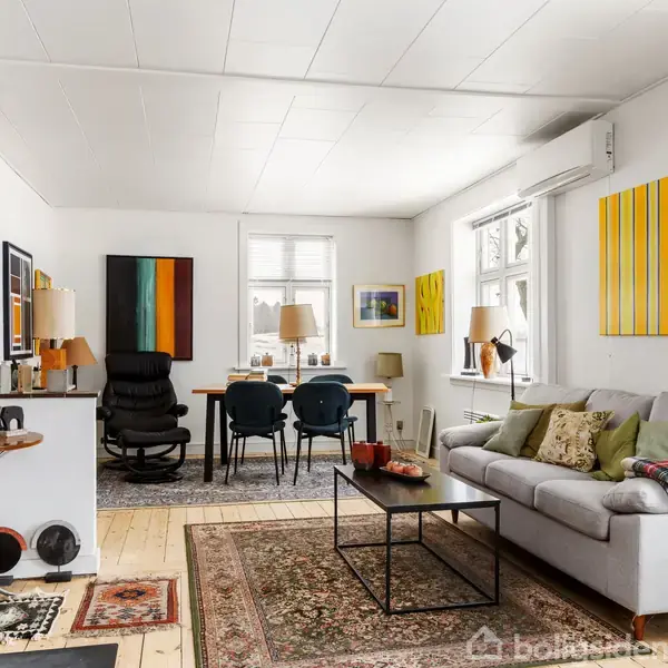Sofa med puder står ved sofabord, mens brændeovn brænder i stuen. Væggene er dekoreret med malerier, og spisebord med stole står ved vindue i baggrunden.