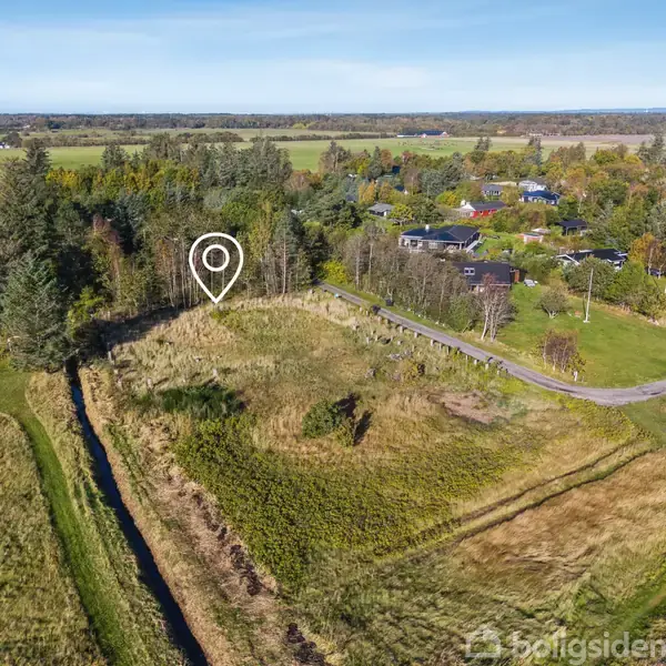 Et stykke åbent græsareal markeret med en hvid indikator, omgivet af skov og huse i en landlig landsby. Markveje og marker er synlige i baggrunden.