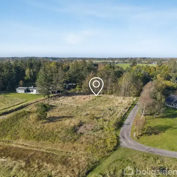 En åben mark med et placermærke i midten, omgivet af en blanding af tæt skov og spredte huse. En snoet vej løber langs markens kant.