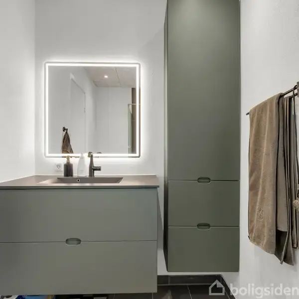 Gråt badeværelsesskab med integreret håndvask står under en vægmonteret firkantet spejl med lyskant. Til højre hænger håndklæder på stænger. Vægoverfladerne er hvide.