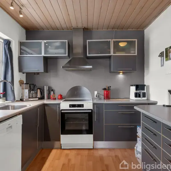 Moderne køkken med kogeplade og ovn i centrum, omgivet af grå skabe og skuffer. Lyset kommer fra loftslamper, og et vindue til venstre bringer naturligt lys ind. Bordplader er lyse med forskellige apparater som elkedel og kaffemaskine placeret på dem.