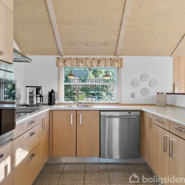 Et moderne køkken med lyst træinventar, indeholder ovn, opvaskemaskine og kaffemaskine. Det er i et solbelyst rum med træloft og vindue, der giver udsigt til et grønt område.