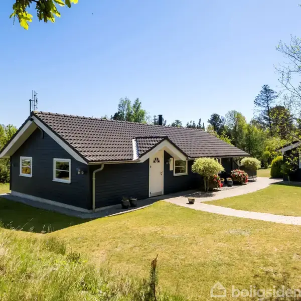 Et sortmalet hus med brun tagstenstak står omkranset af en grøn have, med plæner og buske. En lille sti fører til hoveddøren. Skoven er synlig i baggrunden.